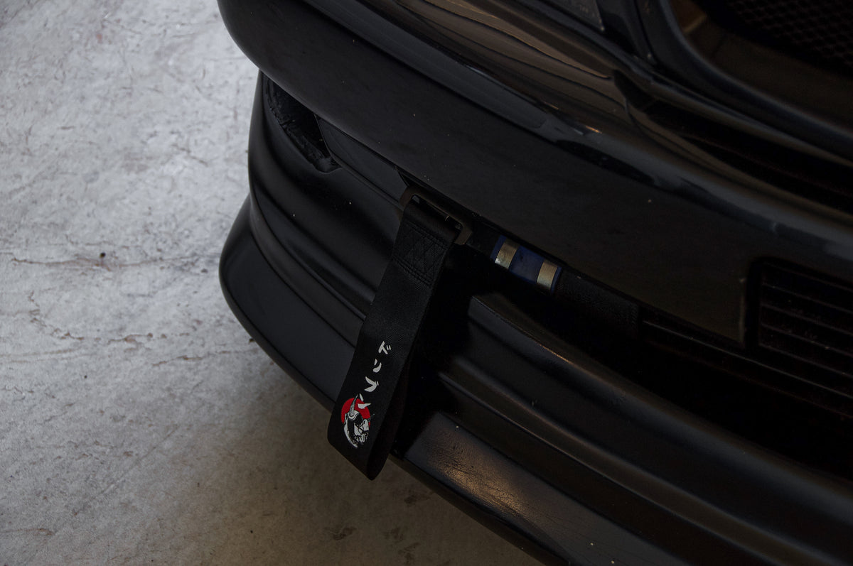 Close-up of the front end of a black car, showcasing an Incompletegl Red Sun Oni Mask Tow Strap with Japanese characters attached to the bumper. The vibrant blue strap extends from the lower grille area to the exterior, and the car is parked on a light-colored concrete surface, highlighting its nod to Japanese culture.