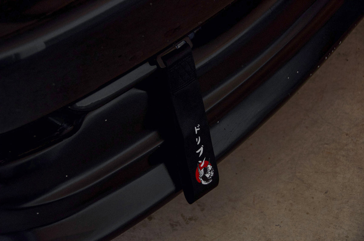 A close-up of a black car grille showcases the Red Sun Oni Mask tow strap from Incompletegl, featuring an illustration of a skull with a helmet. Adorned with Japanese characters, the strap evokes elements of Japanese culture. The car is parked on a concrete surface.