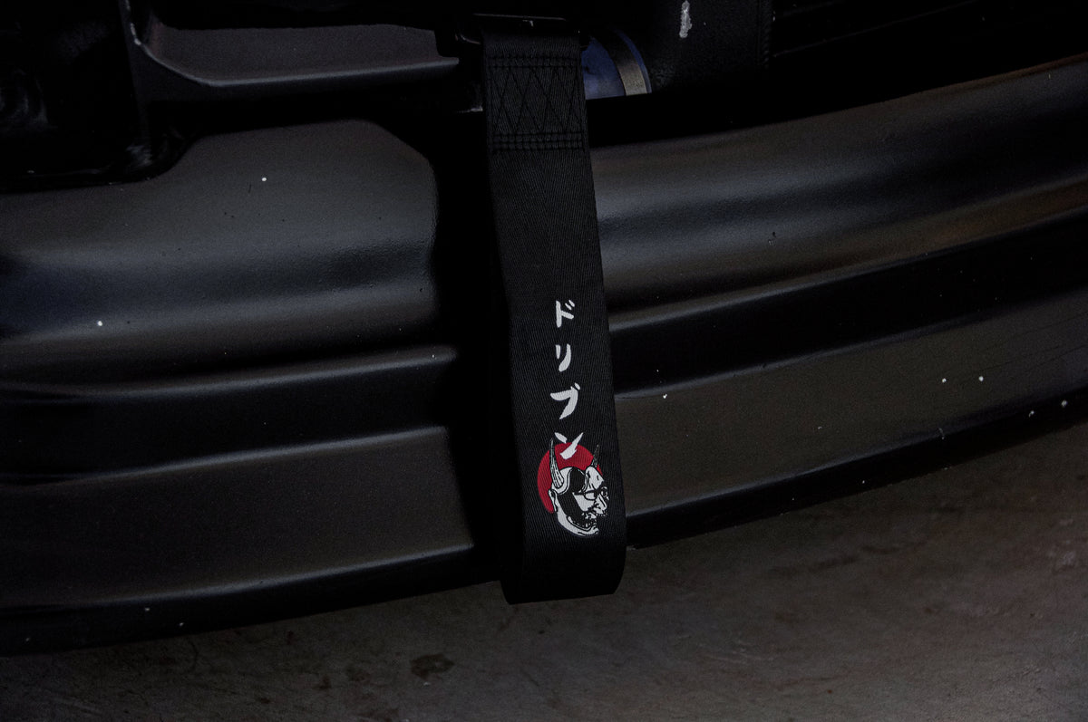 Close-up of a black tow strap, the Red Sun Oni Mask | Tow Strap by Incompletegl, attached to the front bumper. The strap features an illustration of a red and white demon-like face with horns, reminiscent of a traditional Japanese Oni mask. The text "ドリブン" (Japanese for "driven") is printed in white. The car and garage floor are dimly lit, evoking Japanese culture.