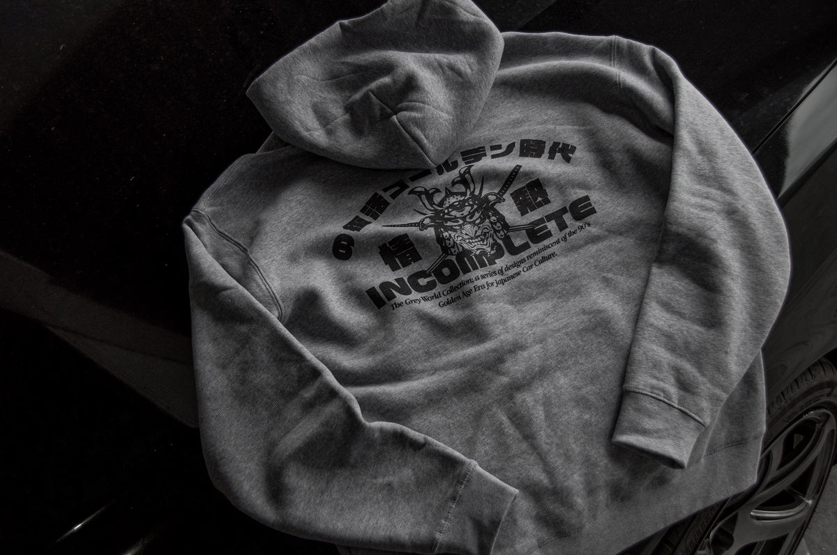 A piece from Incompletegl's Grey World Collection, this grey zip hoodie is laid flat on a black surface. The back features a black graphic design with stylized text in an undecipherable script and English, prominently displaying the word "INCOMPLETE." The design pays homage to the 90's Golden Age Era and includes an intricate illustration.