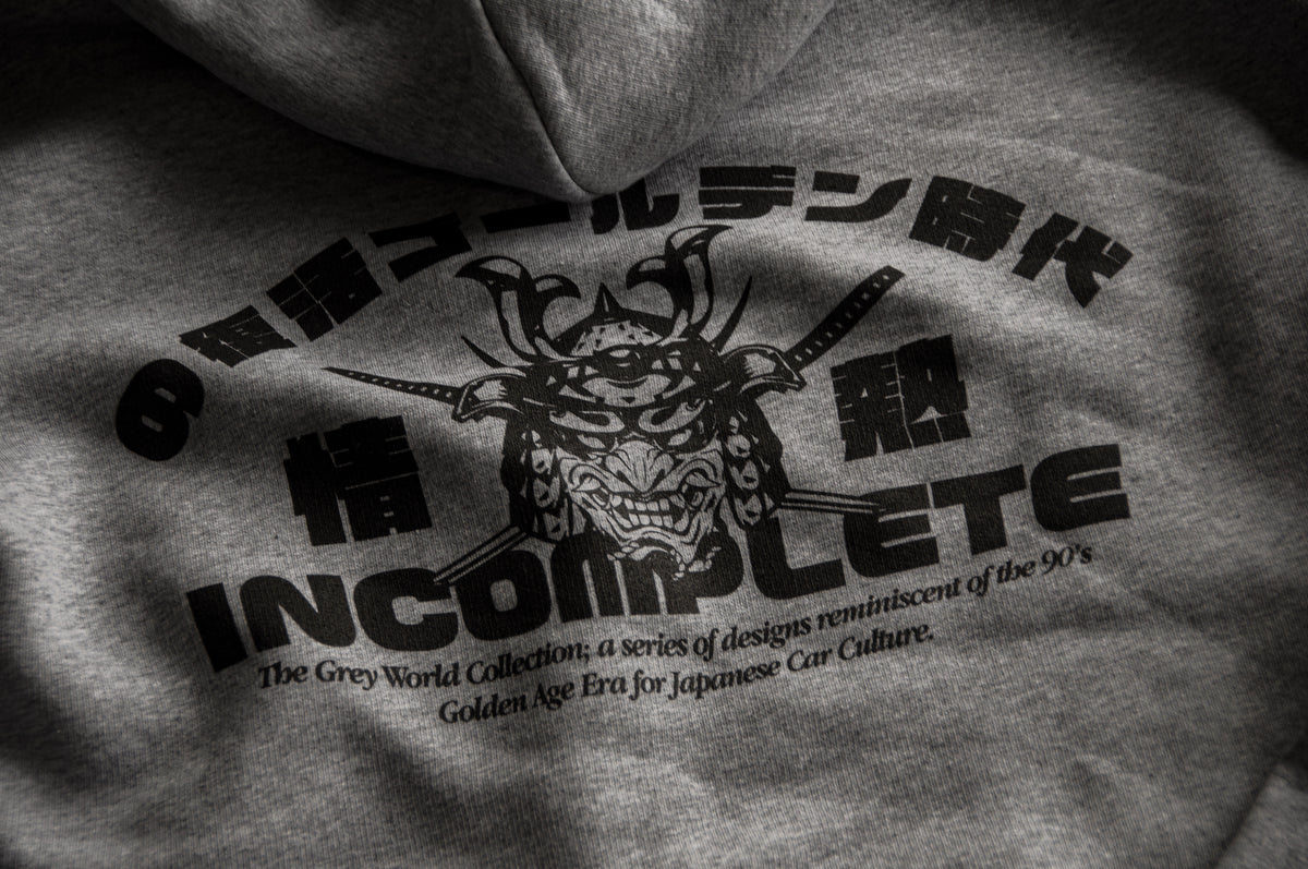 A close-up of the Incompletegl Unisex GREY WORLD! Zip Hoodie in grey from the Grey World Collection showcases a graphic design featuring a samurai helmet with crossed swords behind it. Encircling the helmet are Japanese characters, and beneath it is the text "INCOMPLETE," accompanied by a smaller tagline referencing 90's Japanese Car Culture.