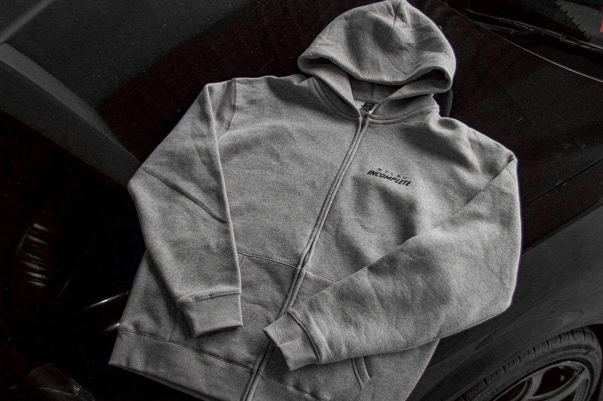 The Incompletegl Unisex GREY WORLD! Zip Hoodie in grey, featuring a small embroidered design on the chest and complete with a hood and pockets, is laid out on the flat surface of a car from the 90's Golden Age Era. This hoodie is made from 320 GSM fabric and available in sizes S to XXL.