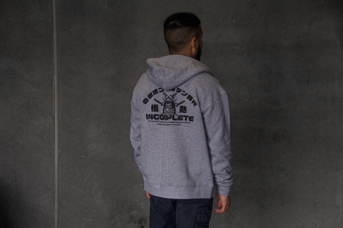 A man with short hair stands with his back to the camera, wearing an Incompletegl Unisex GREY WORLD! Zip Hoodie in gray, which features black text and an image of a house on the back. The background is a plain, dark wall.
