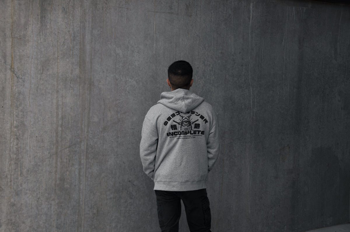 A person with short hair is standing with their back to the camera, wearing an Incompletegl Unisex GREY WORLD! Zip Hoodie in light gray, which features black graphic text and symbols. They are facing a large, plain concrete wall. The overall tone of the image is muted and minimalistic.