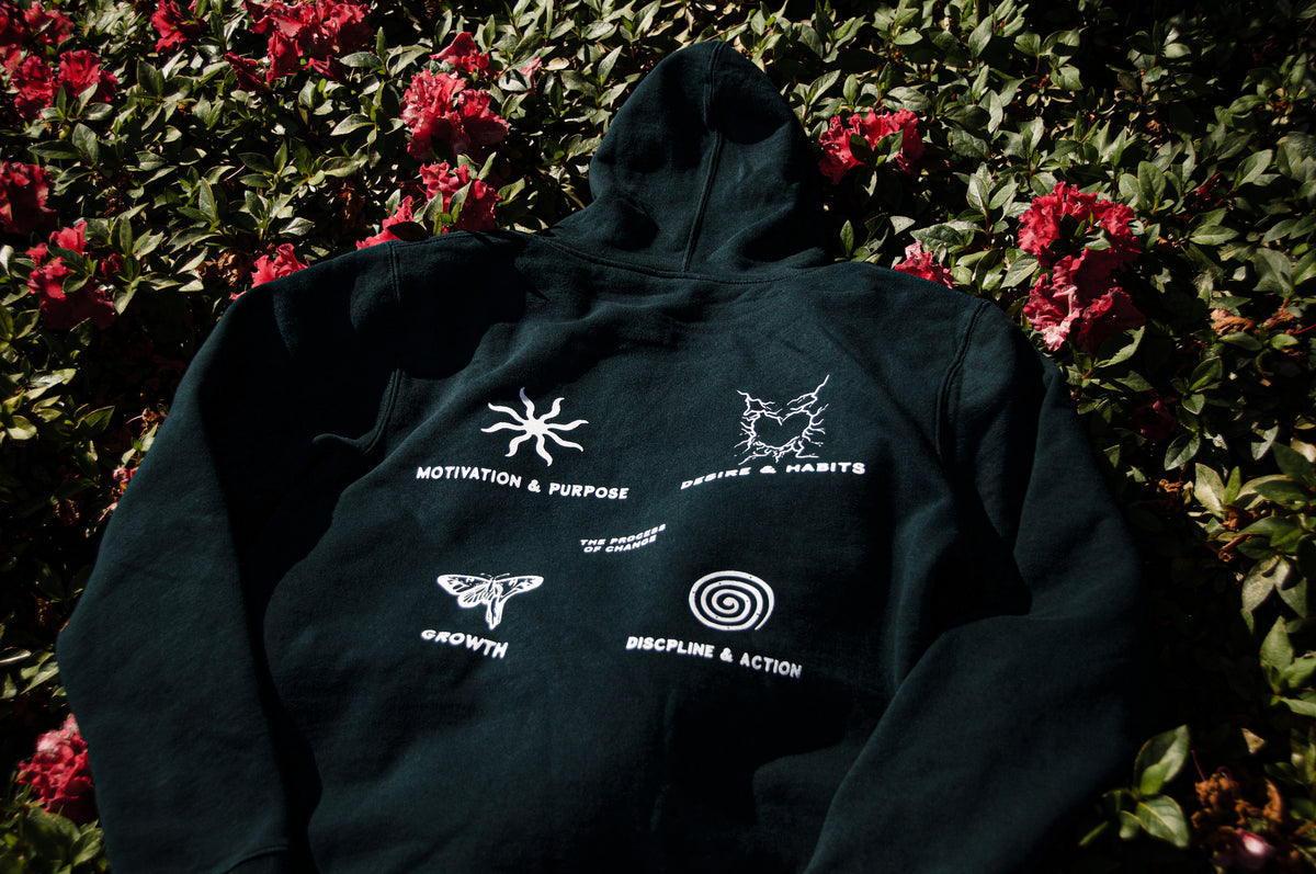 The Motivational OG Hoodie by Incompletegl, a dark green hooded sweatshirt perfect for high-quality hoodie enthusiasts, is laid flat on blooming pink flowers. Various white designs are printed on the sweatshirt, including motifs of a sun, a butterfly, a spiral, and phrases like "MOTIVATION & PURPOSE," "GROWTH," "DISCIPLINE & ACTION," and "HABITS & PURPOSE.