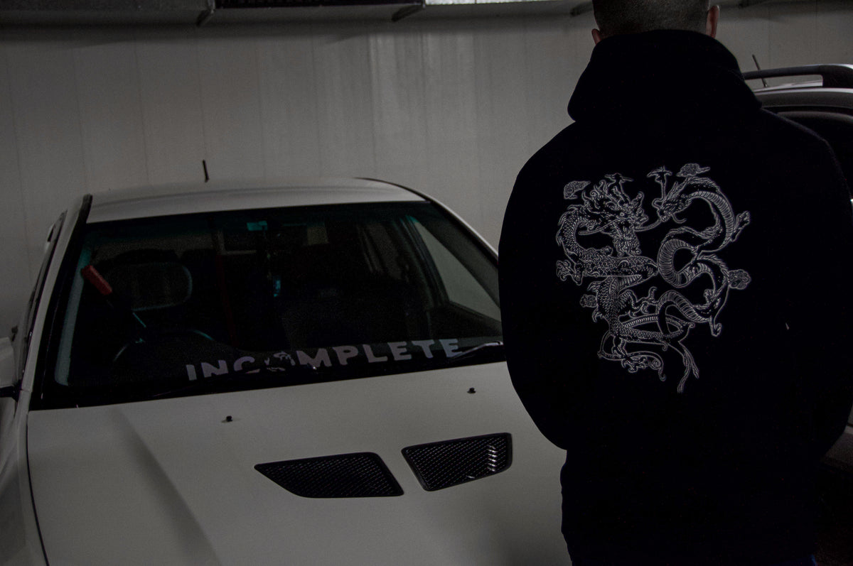 A person wearing an Incompletegl Unisex The Clash of Dragons! Hoodie in black, featuring an intricate Twin Dragons design on the back, stands next to a white car with a vented hood. The car has a large "INCOMPLETE" decal on the inside of the front windshield. The scene appears to be inside a dimly lit garage.