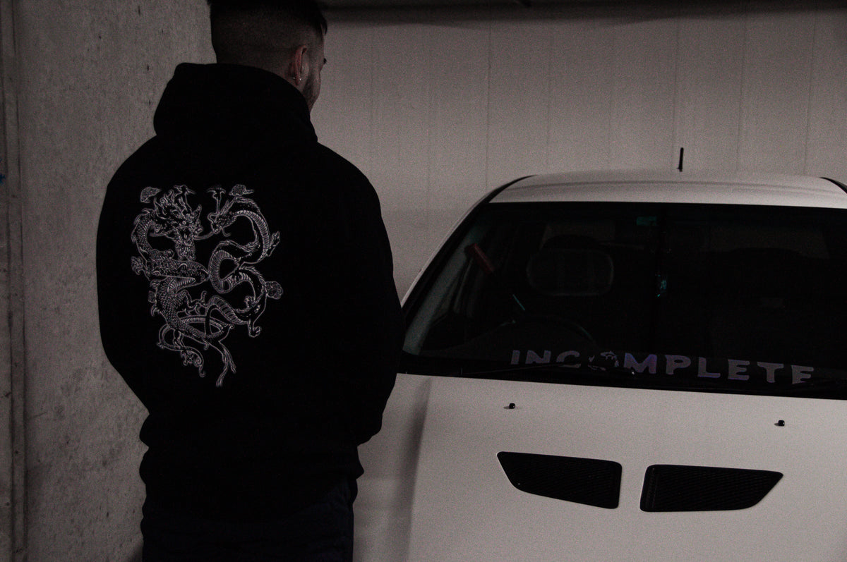 A person wearing an Incompletegl Unisex The Clash of Dragons! hoodie in black, which features an intricate white design on the back, is standing next to a white car. The word "INCOMPLETE" is visible on the windshield of the car. The image is taken in a dimly lit indoor setting.