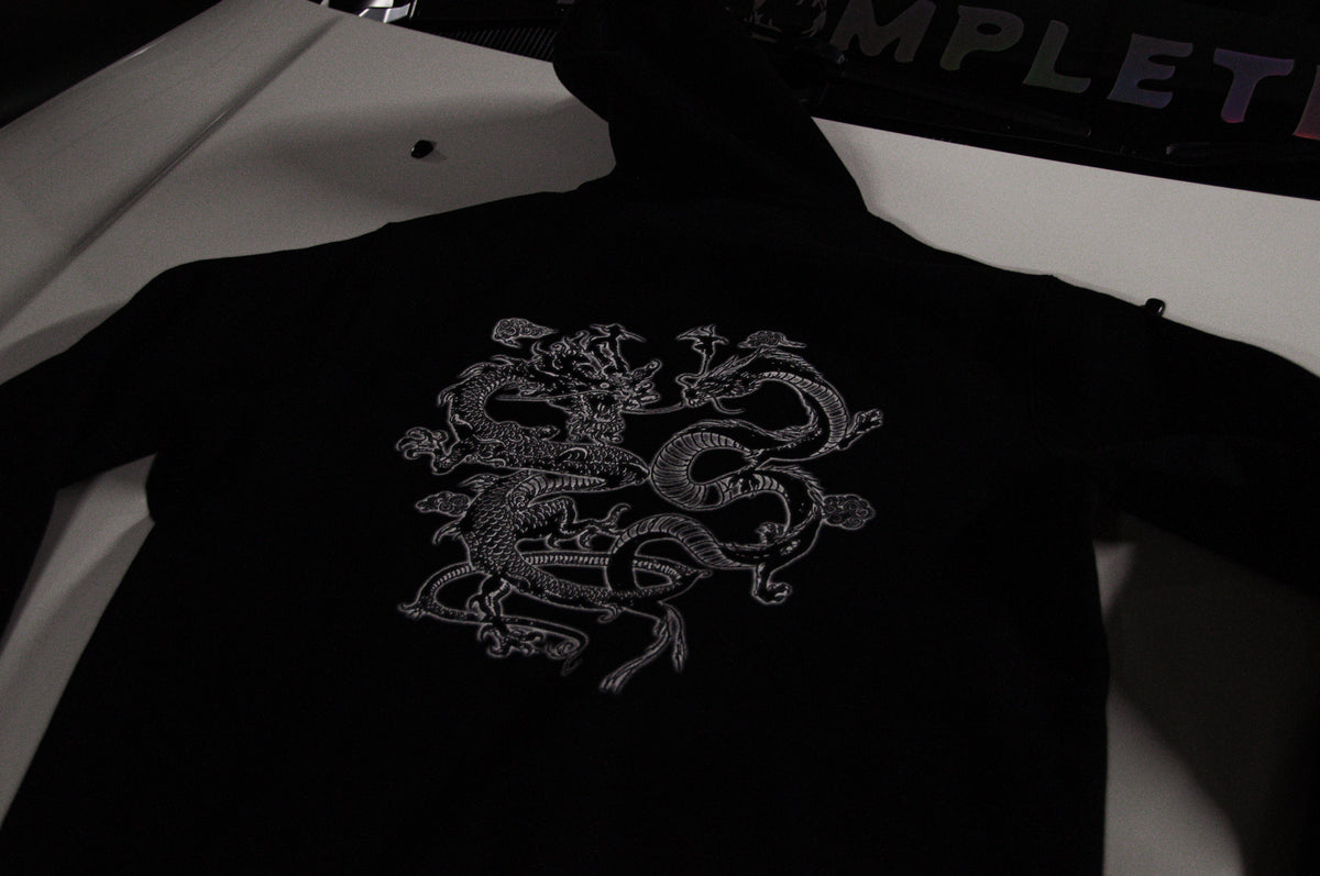 The Incompletegl Unisex The Clash of Dragons! Hoodie in black, made from 300 GSM fabric, features a minimalist artwork of an intricate white embroidered design showcasing Twin Dragons laying flat on a surface. Available in sizes S, M, L, XL, and XXL.