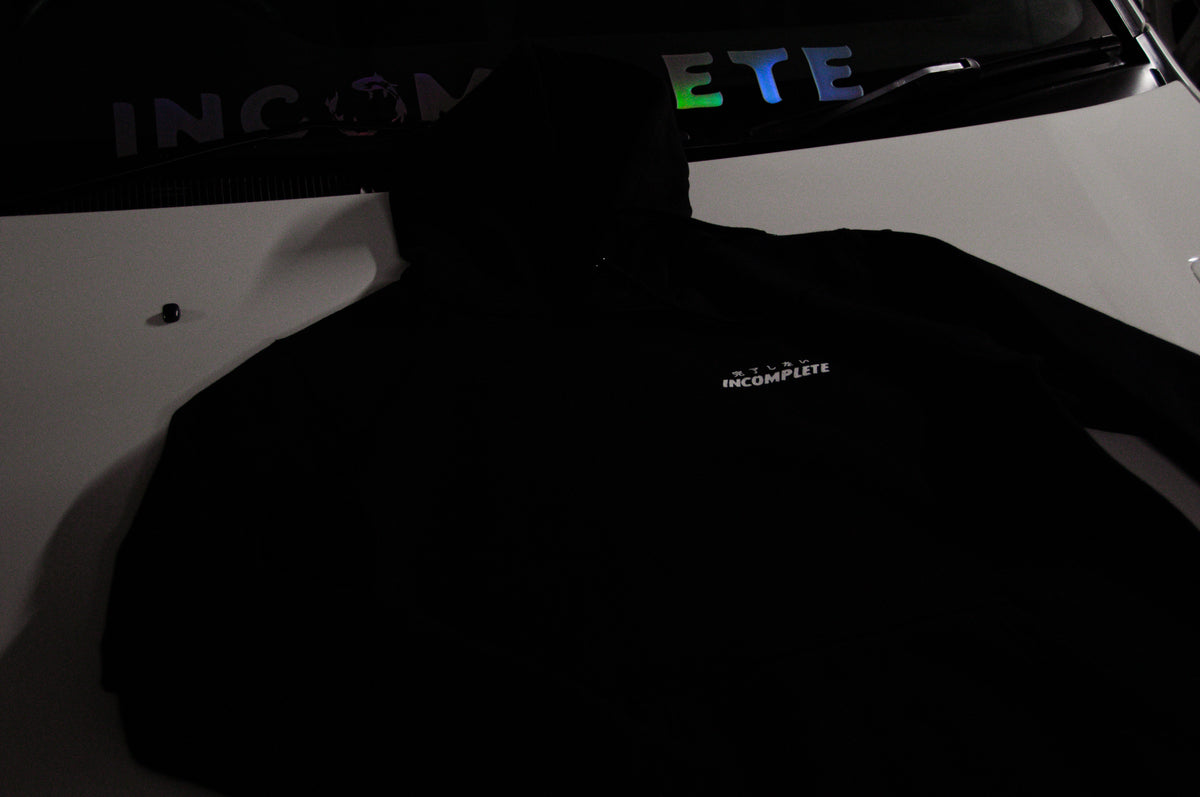 An "Incompletegl" black hoodie from the "Clash of Dragons!" collection is draped over the hood of a white car, featuring the word "INCOMPLETE" printed in white. The car's windshield displays "INCOMPLETE" again in large, colorful letters. The dim lighting adds a moody atmosphere to the scene.