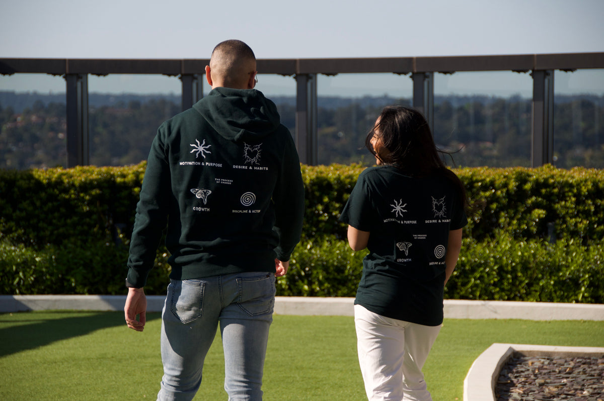 Two people walking on grass with their backs to the camera, both wearing dark green Motivational OG Shirts from Incompletegl that bear white graphic designs and "A Story to Tell" text on the back. A metal fence and shrubbery set the scene, with a scenic landscape unfolding under a clear sky.