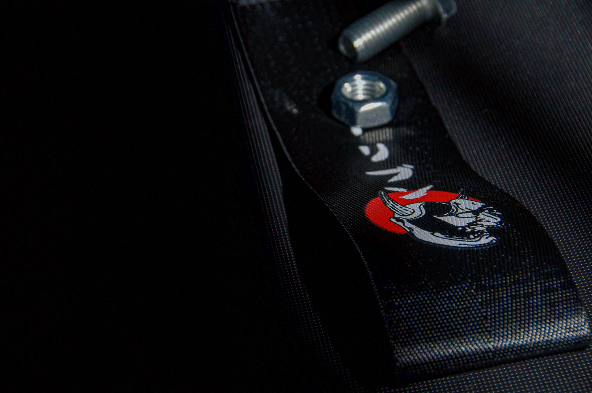 Close-up of the Black "Red Sun Oni Mask" Tow Strap by Incompletegl, featuring a logo of a snarling wolf or dog with striking red and white accents. On the right side of the image, a metal nut and bolt are partially visible, showcasing a design that echoes Japanese culture with elements reminiscent of the Driven Red Sun Oni Mask.