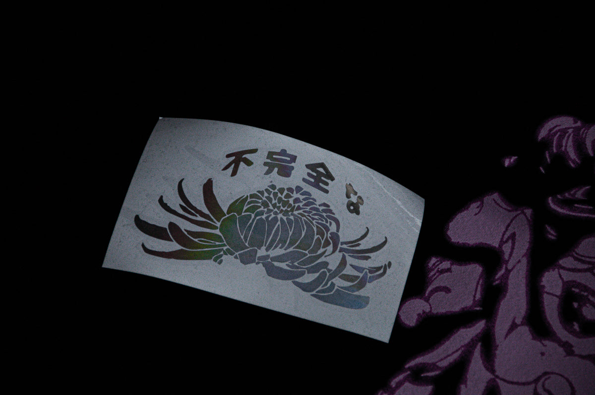 A close-up photograph of an Incompletegl Unisex Undead Geisha x Chrysanthemum T-Shirt in black, featuring Japanese characters above an illustrated chrysanthemum. The flower illustration shows a gradient transitioning from green to purple, set against a dark background that highlights the sticker and the adjacent part of a shaded illustration. Available in sizes S, M, L, XL, and XXL.