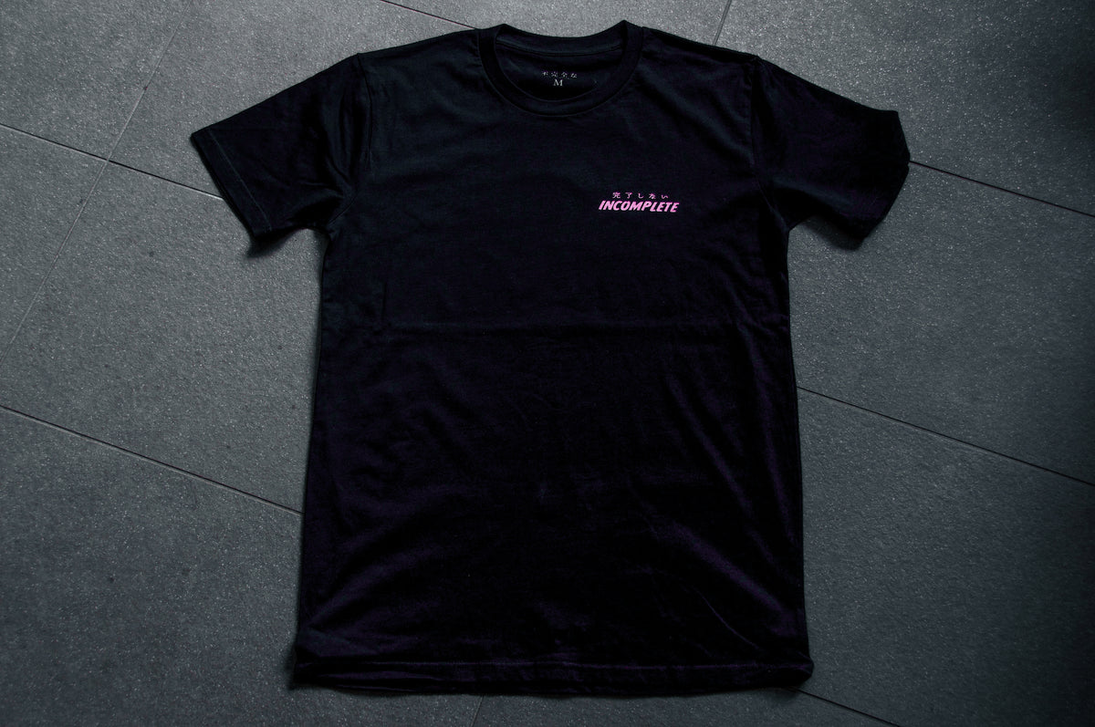 An Incompletegl Unisex Undead Geisha x Chrysanthemum T-Shirt in black, made from 300 GSM material, lies flat on a dark, tiled floor. The shirt features the words "PLEASE RETURN TO" in small, white text, with "INCOMPLETE" in larger, pink text underneath it on the upper left chest area. It pairs perfectly with a stylish hoodie for a modern look. Available in sizes S to XXL.