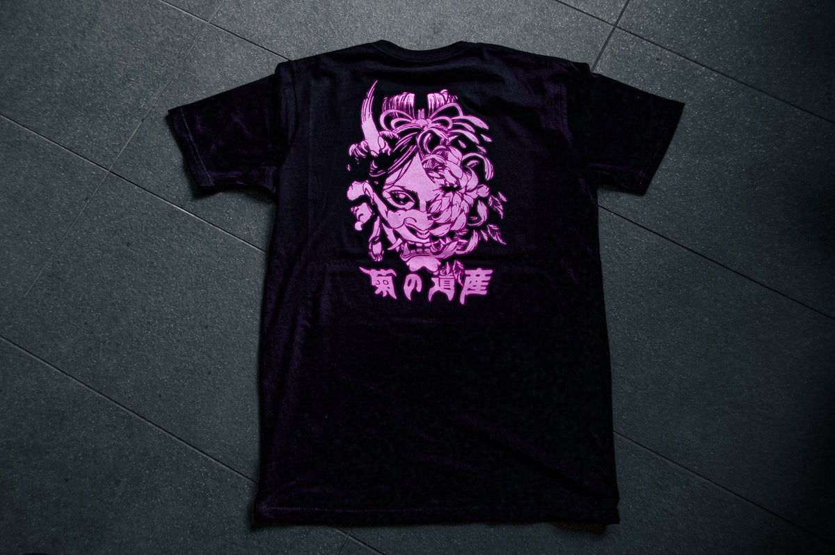 The Incompletegl Unisex Undead Geisha x Chrysanthemum T-Shirt in black features a large back print showcasing a stylized pink chrysanthemum alongside an intricate face adorned with ornamental patterns. Below the design, white Japanese characters add to its cultural elegance. The shirt, laid flat on a dark tiled surface, exudes both sophistication and heritage effortlessly. Available in sizes S, M, L, XL, and XXL and made with 300 GSM fabric.