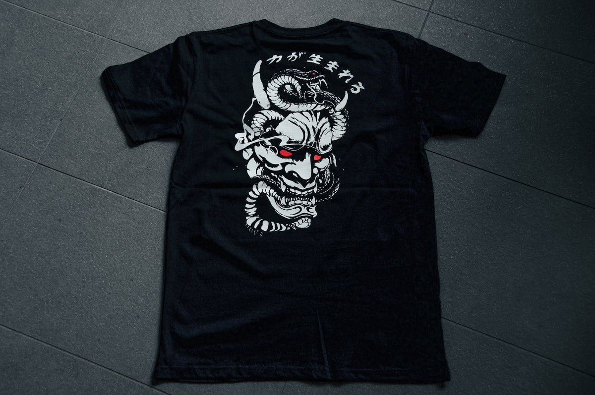 The Incompletegl Unisex Snake x Oni Mask - T-Shirt in black, made from 300 GSM fabric, lays flat on a dark tiled surface. The back of the T-shirt showcases a detailed design of a Hannya Mask with red eyes and Japanese text above it. The white design stands out vividly against the black fabric.