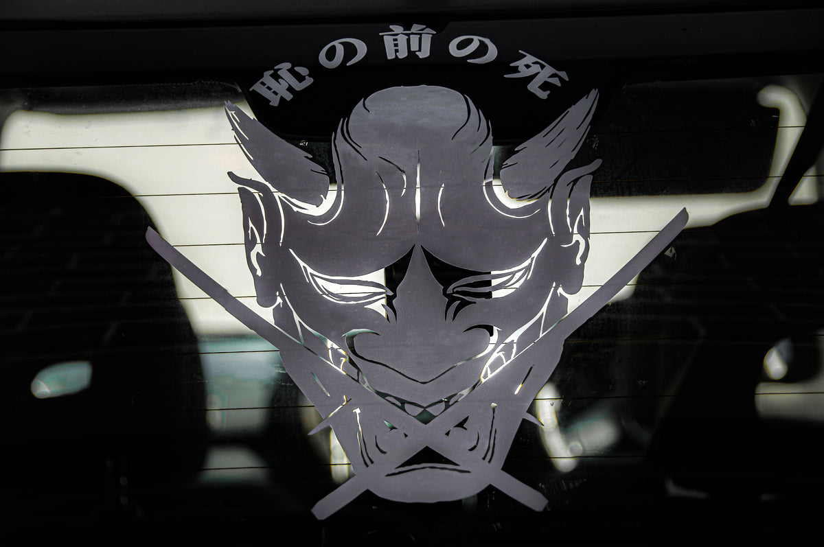Displayed on a glass surface is the Oni Mask "Death before Dishonour" vinyl sticker by Incompletegl, featuring a stylized Oni mask with two horns and a fierce expression. The design includes two crossed katanas beneath the mask and Japanese characters written above, capturing the essence of Japanese culture.