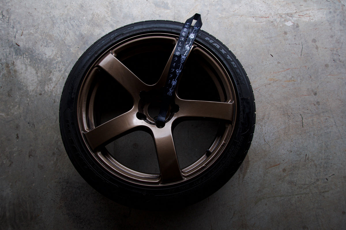 A single bronze-colored, five-spoke car wheel with an attached black tire, positioned vertically against a gray concrete floor. Over one of the spokes, there is "The Mask Collection | Lanyard" by Incompletegl, a high-quality nylon black strap adorned with white Japanese text that reflects aspects of Japanese culture. The lighting creates a contrast with the surrounding shadows.