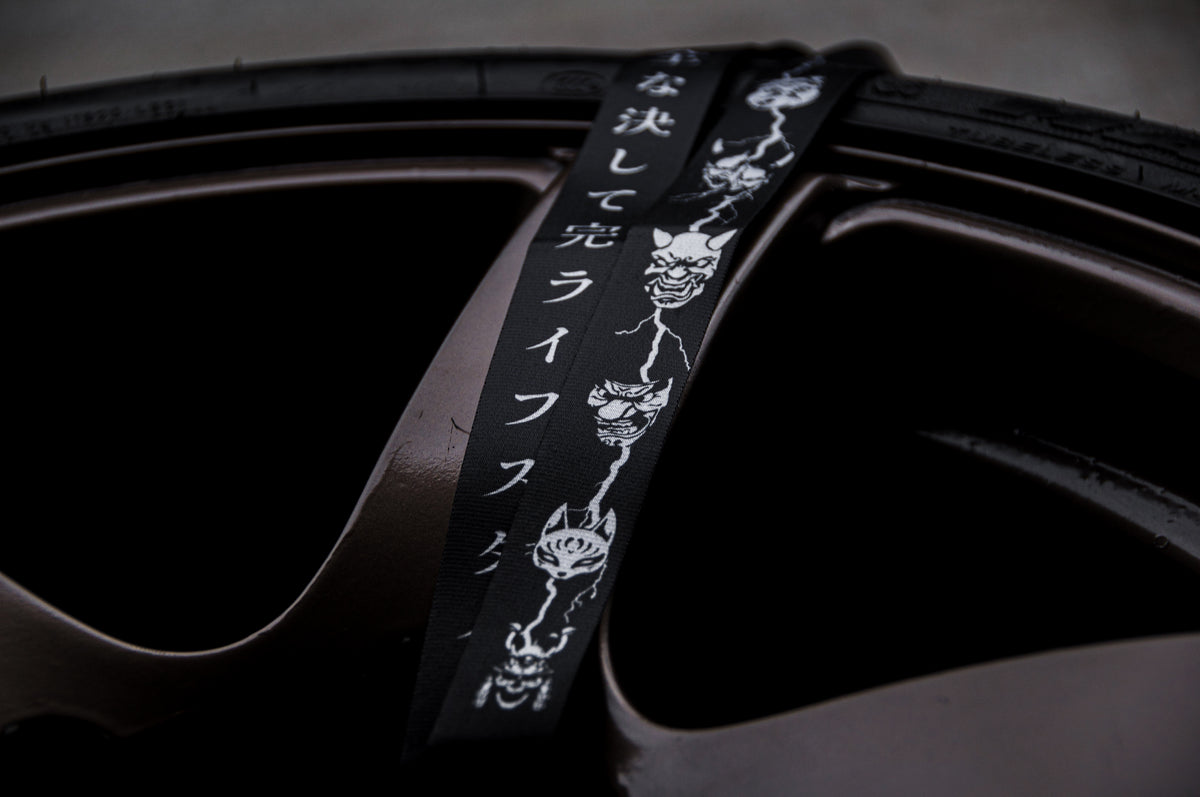 Close-up of a car tire with a black, high-quality nylon strap from The Mask Collection | Lanyard by Incompletegl laid across the rim. The strap features Japanese text and a pattern of white demonic faces, adding a distinctive and edgy style to the image. The tire and rim are dark brown, complementing the automotive lifestyle inspired design.