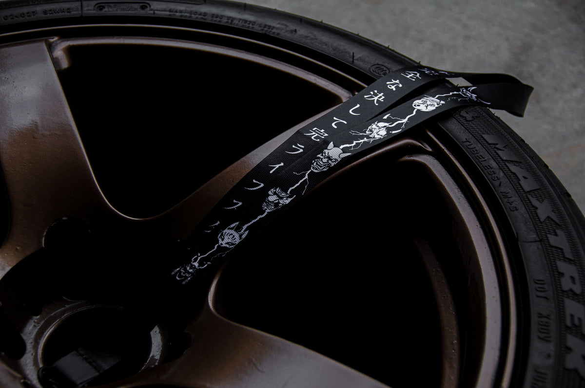 Close-up of a bronze-colored car wheel with The Mask Collection | Lanyard by Incompletegl, adorned in a striking black and white floral pattern, securely attached. The image highlights the intricate design of the high-quality nylon lanyard and the sleek finish of the wheel. The tire brand "Maxxis" is partially visible on the sidewall, embodying an automotive lifestyle.