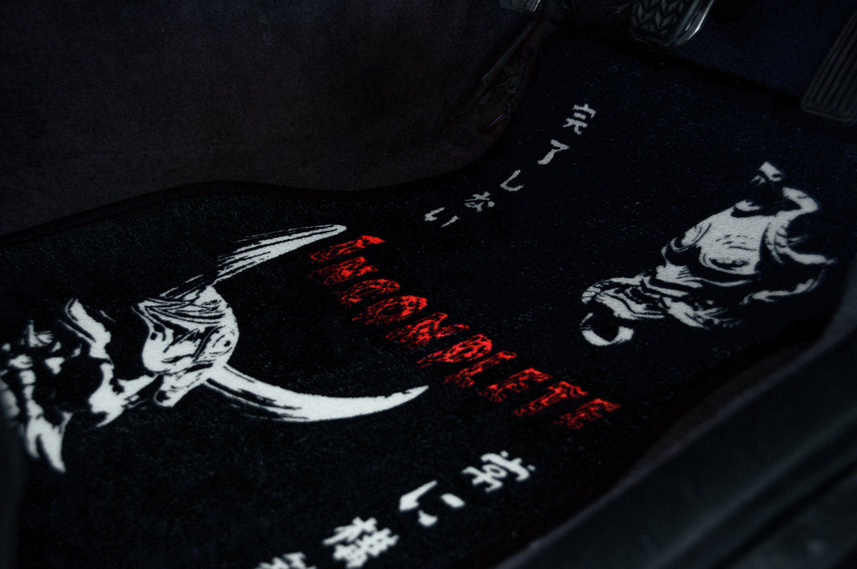 A close-up of the "The Oni Masks | Floor Mats" from Incompletegl displays a black car mat adorned with striking red and white illustrations of a menacing skull and an evocative horned figure reminiscent of traditional Oni masks. It also features Japanese characters and the word "INCOMPLETE" boldly presented in red letters.