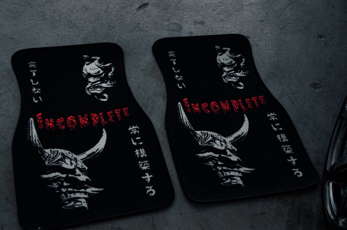 Two black floor mats from Incompletegl, adorned with intricate oni mask designs and Japanese text. The central English text reads "Incomplete" in bold red letters. These mats, capturing a strong Japanese spirit, are placed on a metallic surface.