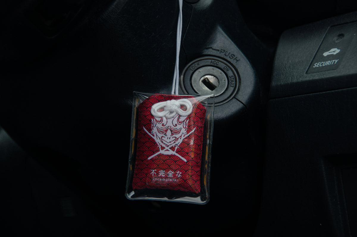 An Oni Mask Omamori Charm | Fearless! by Incompletegl, featuring a red and white fearless oni mask design with intricate patterns enclosed in a plastic case, hangs from a car's ignition switch.