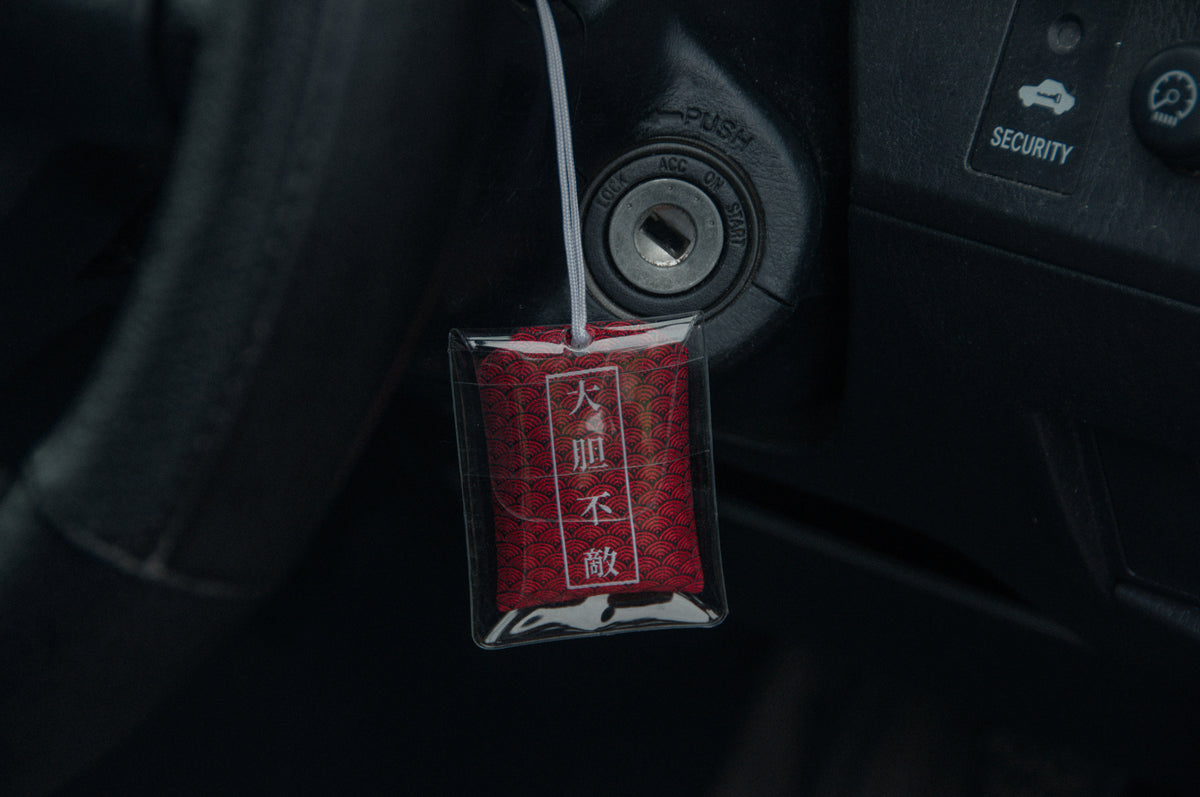 An Oni Mask Omamori Charm from Incompletegl, emblazoned with Japanese characters, hangs from a keyhole in a car. Encased in a clear plastic cover and suspended by a white string, the Fearless! amulet sways gently against the dark vehicle dashboard adorned with buttons labeled "PUSH" and "SECURITY".