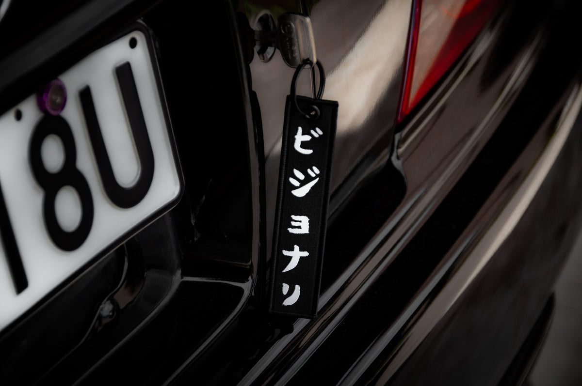 Close-up of a car's rear, featuring a license plate reading "18U" and an Incompletegl "The Samurai & His Vision | Jet Tag" keychain with white vertical Japanese text hanging from the trunk—a nod to Japan's most iconic symbols.