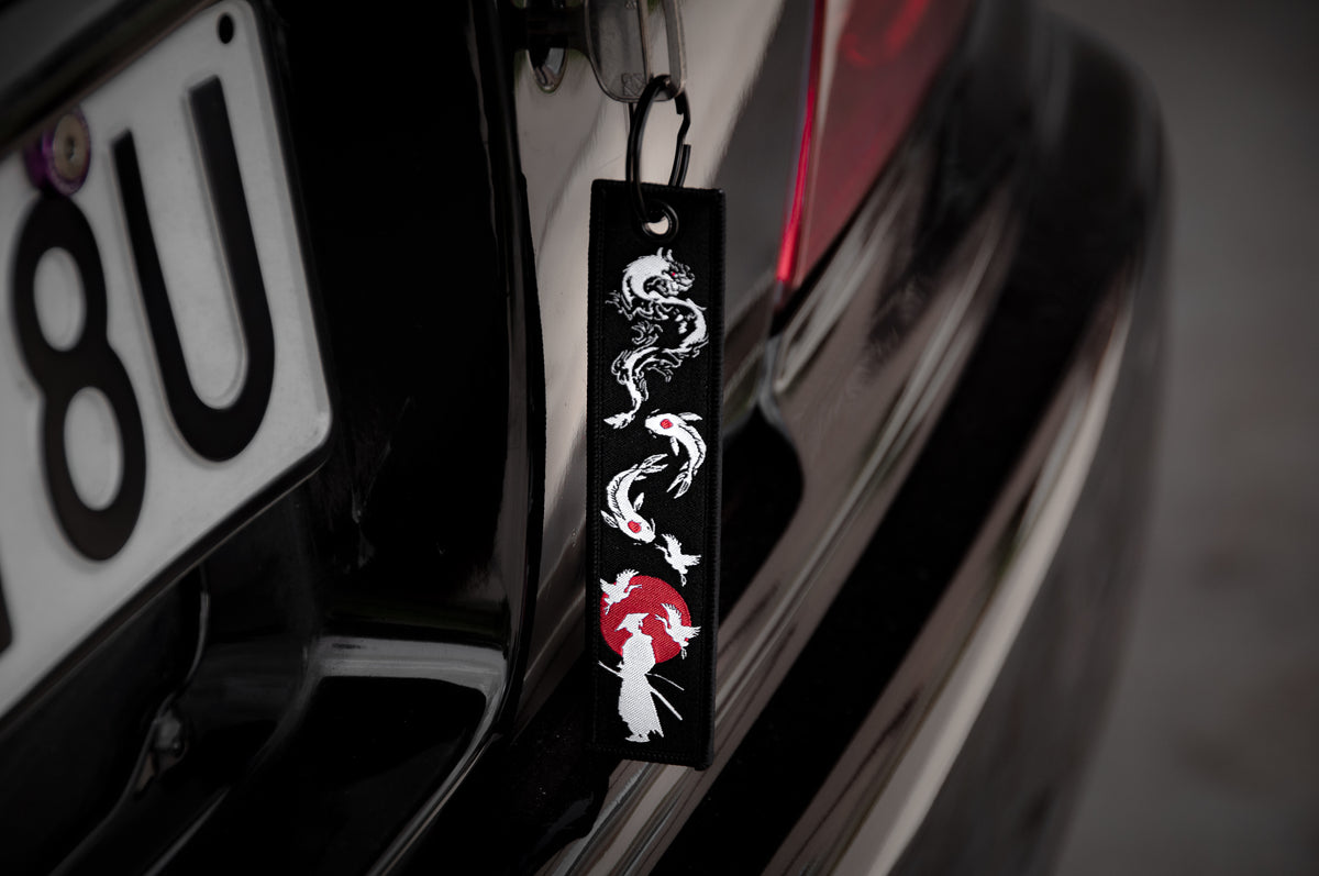 A close-up of a car with "The Samurai & His Vision" Jet Tag by Incompletegl hanging near the license plate. The black fabric strip is embroidered with a white and red dragon and a figure holding an umbrella, echoing Japan's most iconic symbols.