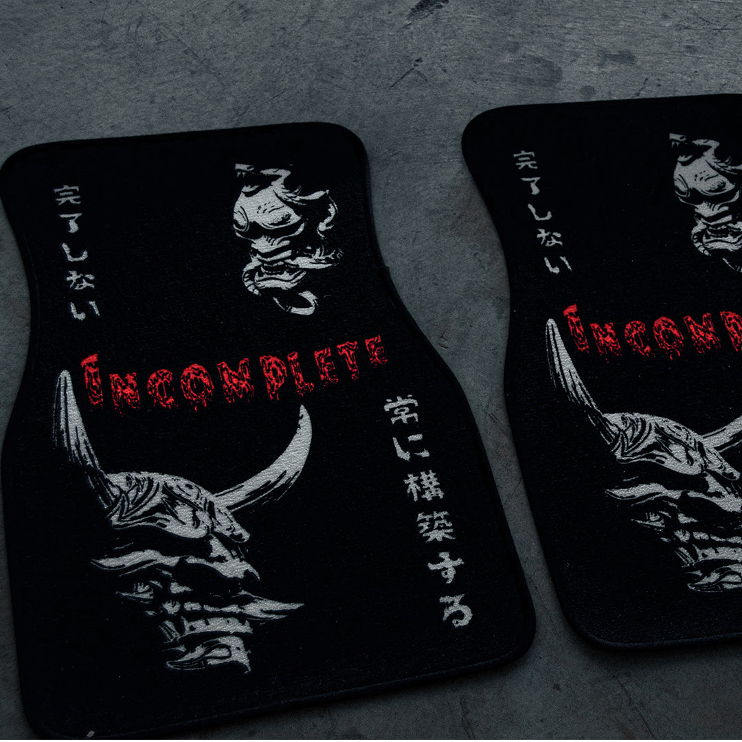 Two black car floor mats feature a stylized, menacing face with horns reminiscent of Oni masks and intricate white Japanese characters. The word "INCOMPLETE" is prominently printed in red, jagged letters across the mats on a textured gray background. These floor mats are part of "The Oni Masks" collection by Incompletegl.