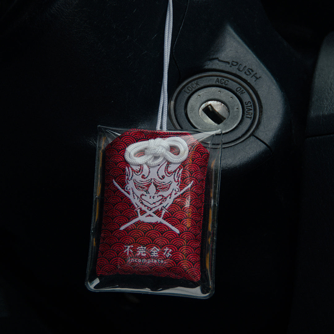 A close-up photo of a car ignition with a key slot features the Oni Mask Omamori Charm | Fearless! by Incompletegl hanging in front. This red Japanese amulet for protection showcases a white oni mask with horns and stylized waves on the fabric. The dark background highlights the fearless details of the charm.