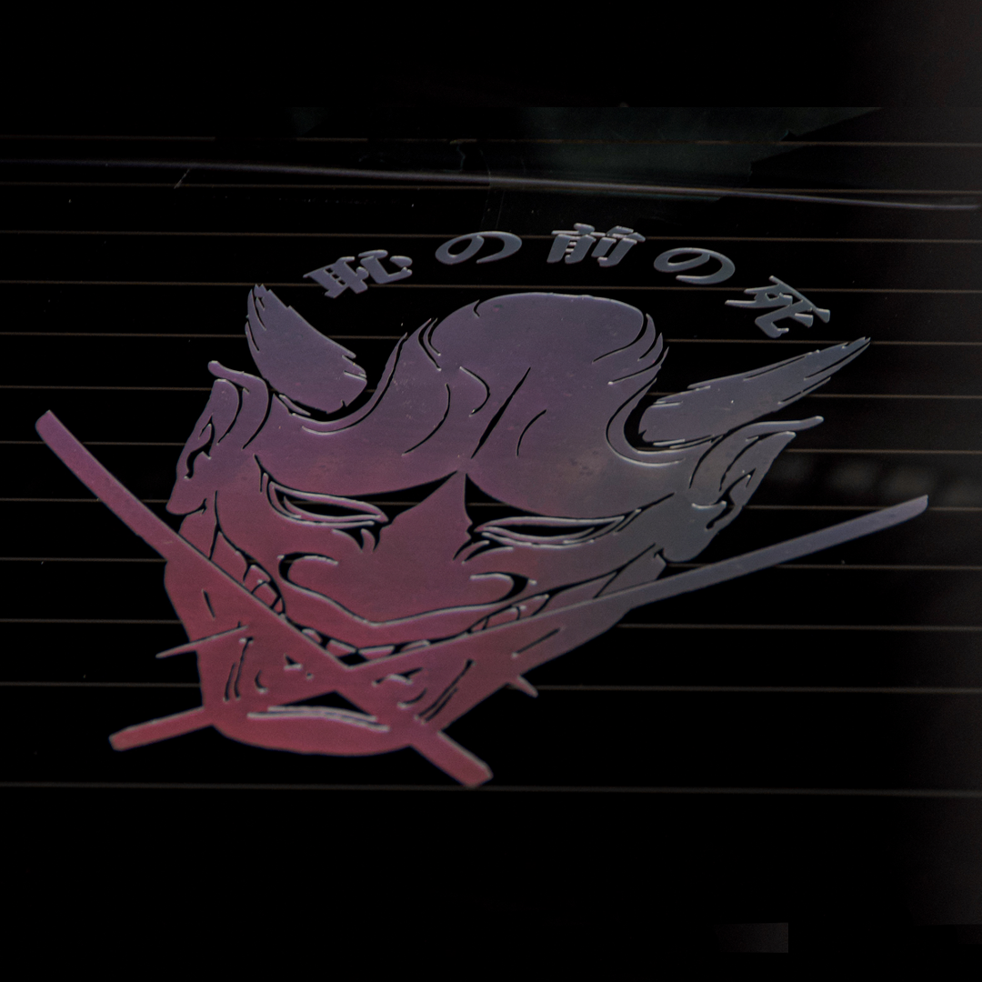 The Oni Mask "Death before Dishonour" Vinyl Sticker by Incompletegl showcases a stylized Japanese oni mask on a dark background, adorned with intricate details like horns and sharp teeth. Japanese characters are visible above the mask, adding to its cultural authenticity. The sticker's metallic, reflective surface shimmers with shades of purple and blue, capturing the essence of Japanese culture beautifully.
