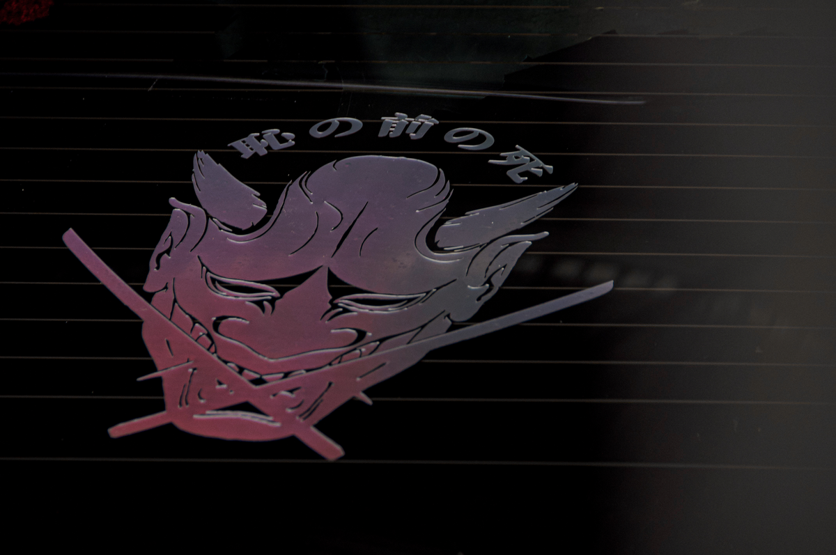The Oni Mask "Death before Dishonour" vinyl sticker by Incompletegl showcases a dark, reflective surface featuring a stylized purple Oni mask with horns and sharp features. Japanese characters hover above the design, while two crossed katana lie below, creating an enigmatic and artistic representation deeply rooted in Japanese culture.