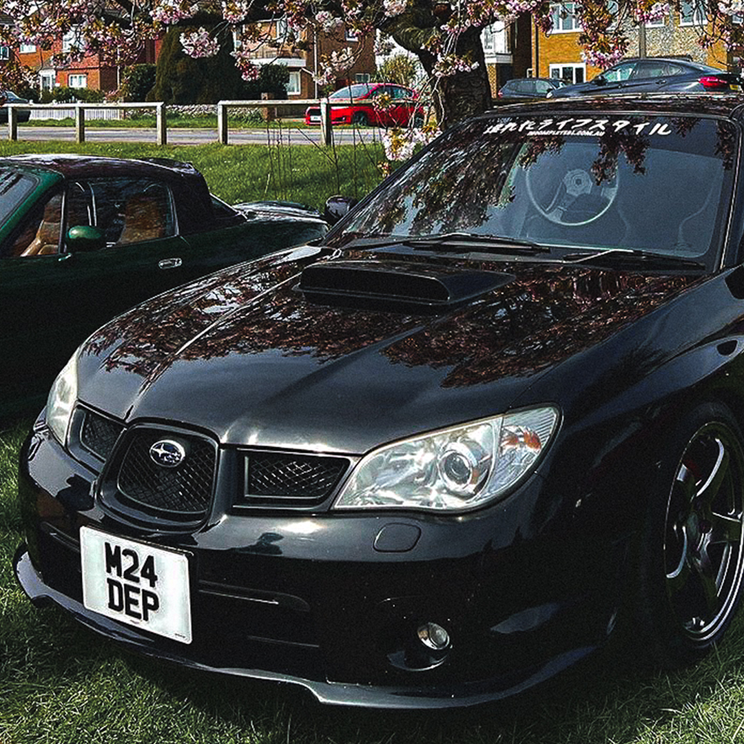 A black Subaru Impreza with a custom hood scoop is parked on a grass field during one of the local car meets. Sporting a license plate reading "M24 DEP" and a large Incompletegl Reverse Style! Car Window Banner - Matte White, Holographic with Japanese characters on the windshield, it's surrounded by other cars and cherry blossom trees in the background.